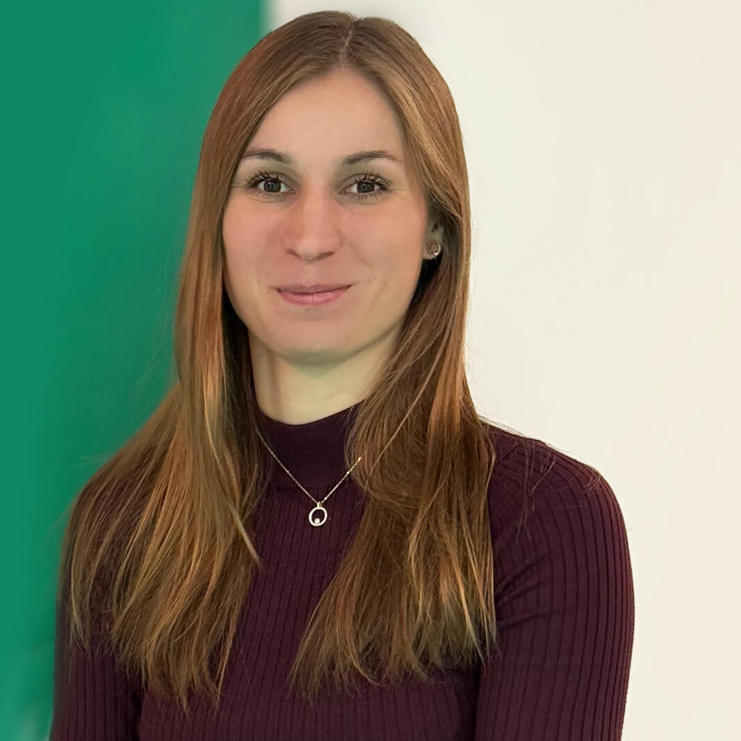 Eine Person mit langem, braunem Haar trägt einen dunkelroten Pullover und eine Kette mit einem kreisförmigen Anhänger. Der Hintergrund ist in den Farben grün und weiß gehalten. Die Person lächelt dezent.