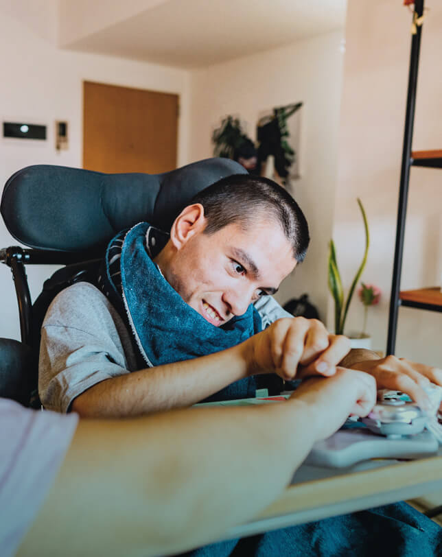 Oskar Intensivpflege Ambulante Intensivpflege Kl Ein lächelnder Mann sitzt in einem Rollstuhl und spielt mit einem Controller. Der Raum ist hell, mit Pflanzen im Hintergrund. Eine andere Person sitzt neben ihm.