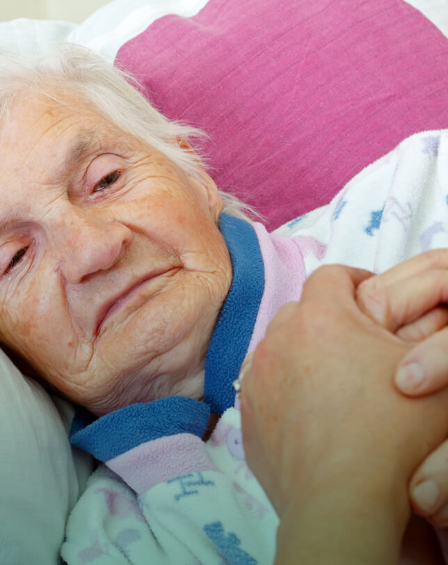 Oskar Intensivpflege Ambulante Intensivpflege Kl Eine ältere Person liegt im Bett auf einem Kopfkissen. Sie trägt ein Schlafanzugoberteil. Eine andere Hand hält sanft die Hand der älteren Person. Der Hintergrund ist teilweise mit einem rosa Kissen bedeckt.