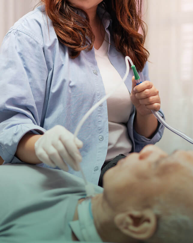 Oskar Intensivpflege Beatmungspflege Tracheostoma Kl Eine Frau in blauer Bluse und weißen Handschuhen hält ein medizinisches Gerät über einen älteren Mann, der liegt. Der Fokus liegt auf dem Gerät, während die Frau im Hintergrund steht.