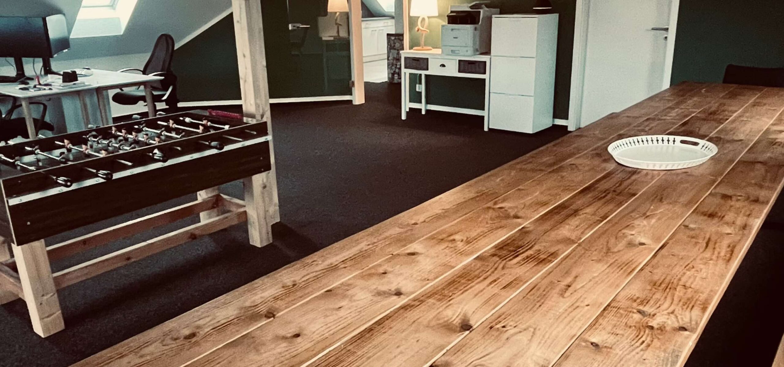 Großer Raum mit Holztisch im Vordergrund, Kicker-Tisch links, Schreibtische mit Bürostühlen hinten, und Regale mit Lampen und Ordnern. Teppichboden und schräge Decke vorhanden.