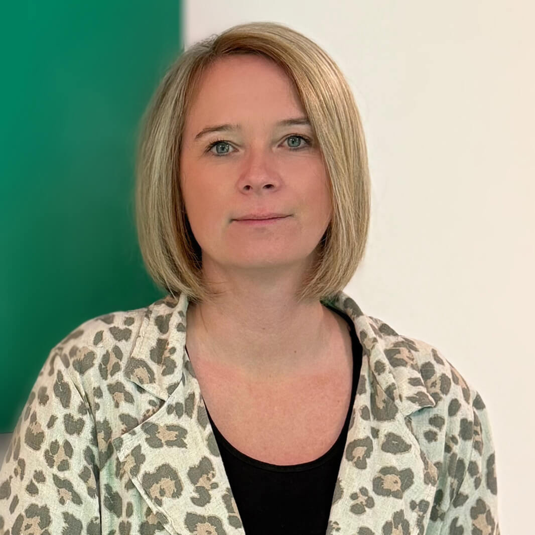 Eine Person mit schulterlangem, blondem Haar steht vor einem Hintergrund, der aus einem grünen und einem weißen Bereich besteht. Sie trägt eine hell gemusterte Bluse und schaut direkt in die Kamera.