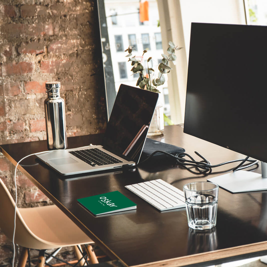 Ein moderner Schreibtisch mit einem Laptop, einem großen Monitor, einer Tastatur und einer Maus. Auf dem Tisch stehen eine Metallflasche, ein Glas Wasser und ein Notizbuch. Im Hintergrund ist eine Ziegelwand sichtbar.