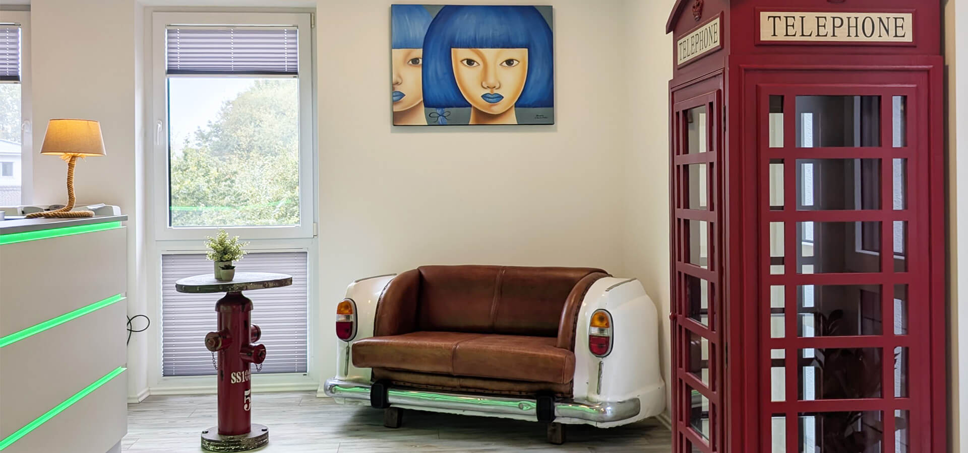 Ein Raum mit einer roten Telefonzelle, einer Couch aus Autoteilen und einem Bild an der Wand. Links ein Empfangstresen mit grünen Leuchten, rechts eine Stehlampe und Fenster mit Jalousien.