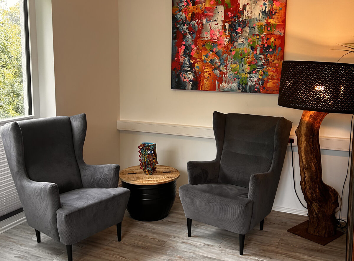 Ein Raum mit zwei grauen Sesseln, einem kleinen runden Tisch dazwischen. Auf dem Tisch steht eine bunte Skulptur. An der Wand hängt ein buntes Gemälde. Eine Lampe mit Holzboden und dunklem Schirm steht rechts.
