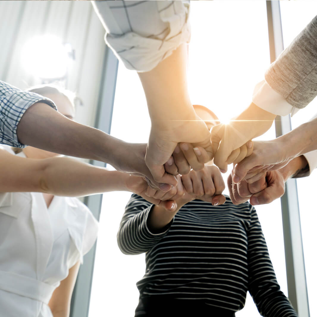 Sechs Personen stehen im Kreis und strecken ihre Fäuste zusammen in die Mitte, als Zeichen von Teamgeist und Zusammenarbeit. Sonnenlicht scheint durch ein Fenster im Hintergrund.