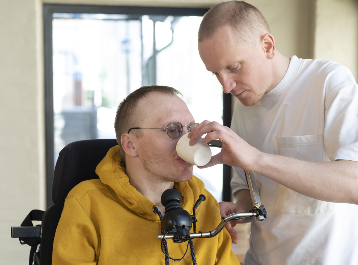 Ein Mann im Rollstuhl mit gelbem Hoodie trinkt aus einem Becher, der ihm von einem Pfleger in weißer Kleidung gereicht wird. Beide befinden sich in einem Raum mit großem Fenster im Hintergrund.