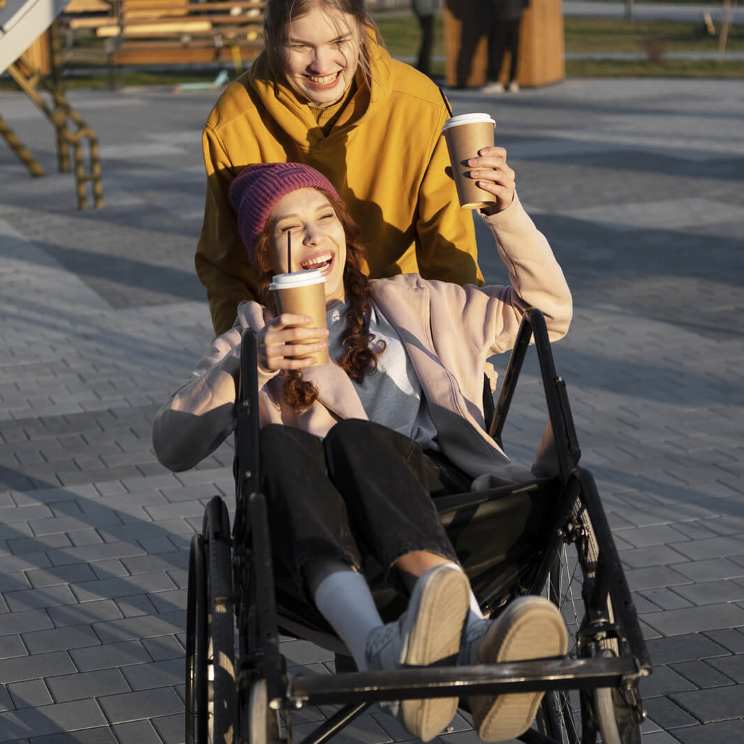 Zwei lachende Personen im Freien. Eine Person im Rollstuhl hält einen Kaffeebecher und trägt eine pinke Mütze. Die andere Person in gelber Jacke steht dahinter und hält ebenfalls einen Kaffeebecher. Sie genießen den sonnigen Tag.