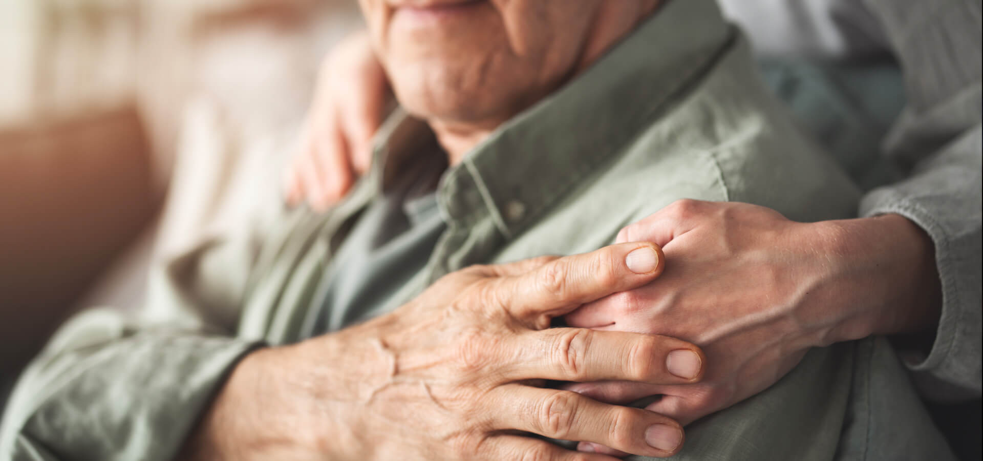Oskar Intensivpflege Home Panorama 1920x900 Ältere Person in Hemd sitzt, während jemand im Hintergrund sanft die Schulter berührt, was Nähe und Unterstützung vermittelt. Hände sind nah beieinander positioniert, der Hintergrund ist unscharf.