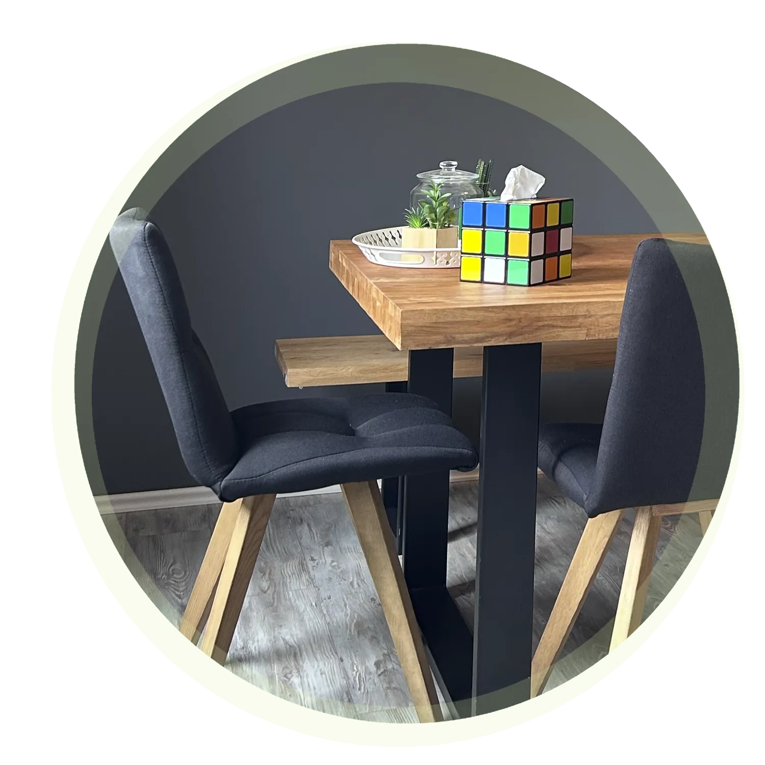 Ein Holztisch mit schwarzem Metallgestell, flankiert von zwei gepolsterten Stühlen. Auf dem Tisch stehen ein Rubik's Cube, eine Glasbox mit Deckel und eine kleine Grünpflanze. Der Boden ist aus grauem Holz.