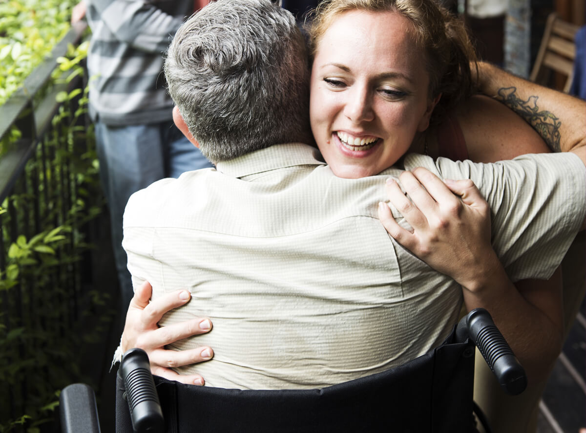 Eine Frau umarmt einen Mann, der in einem Rollstuhl sitzt. Die Frau lächelt fröhlich. Im Hintergrund sind unscharf Pflanzen und eine weitere Person zu sehen.