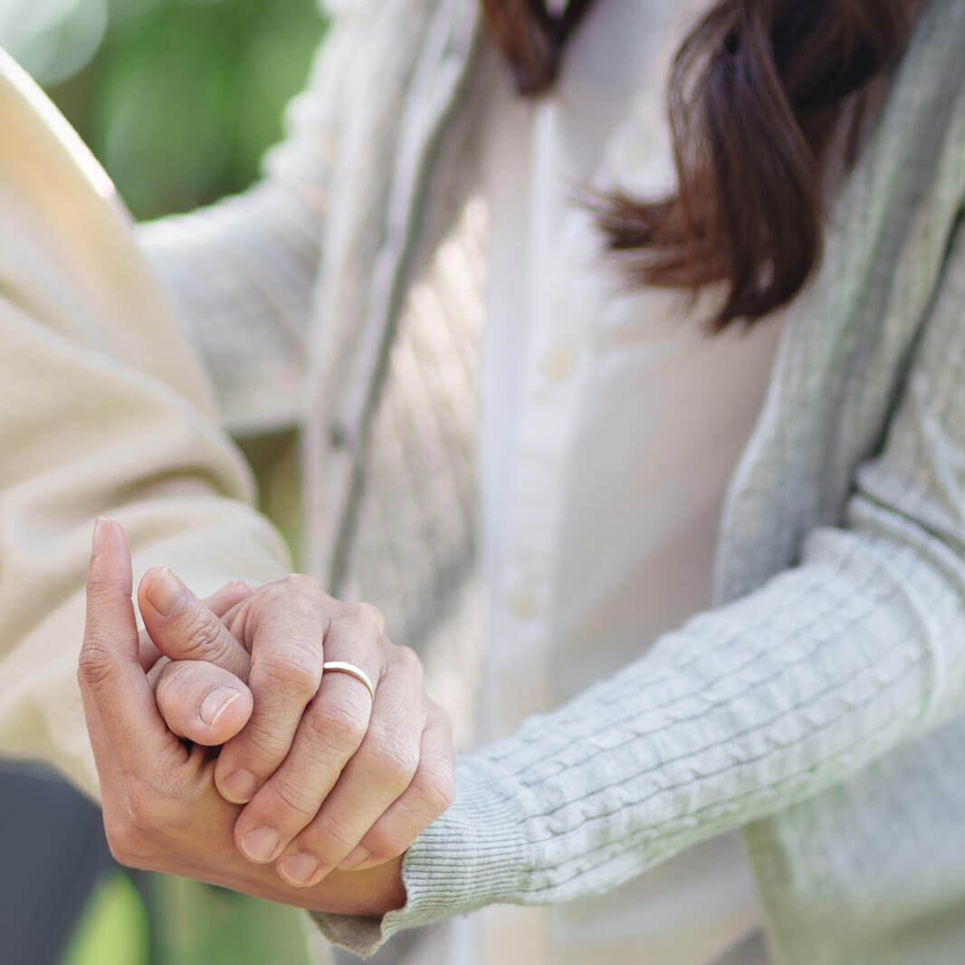 Zwei Personen halten sich sanft an den Händen. Der Fokus liegt auf den Händen; der Hintergrund ist unscharf. Beide tragen helle Kleidung, und eine Person trägt einen grauen Strickcardigan.