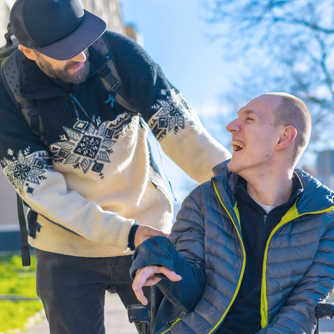 Oskar Intensivpflege Pflege Campus Ein Mann im Rollstuhl lächelt einen anderen Mann an, der einen schwarzen Hut und eine Strickjacke mit Schneeflockenmuster trägt. Sie befinden sich im Freien an einem sonnigen Tag.