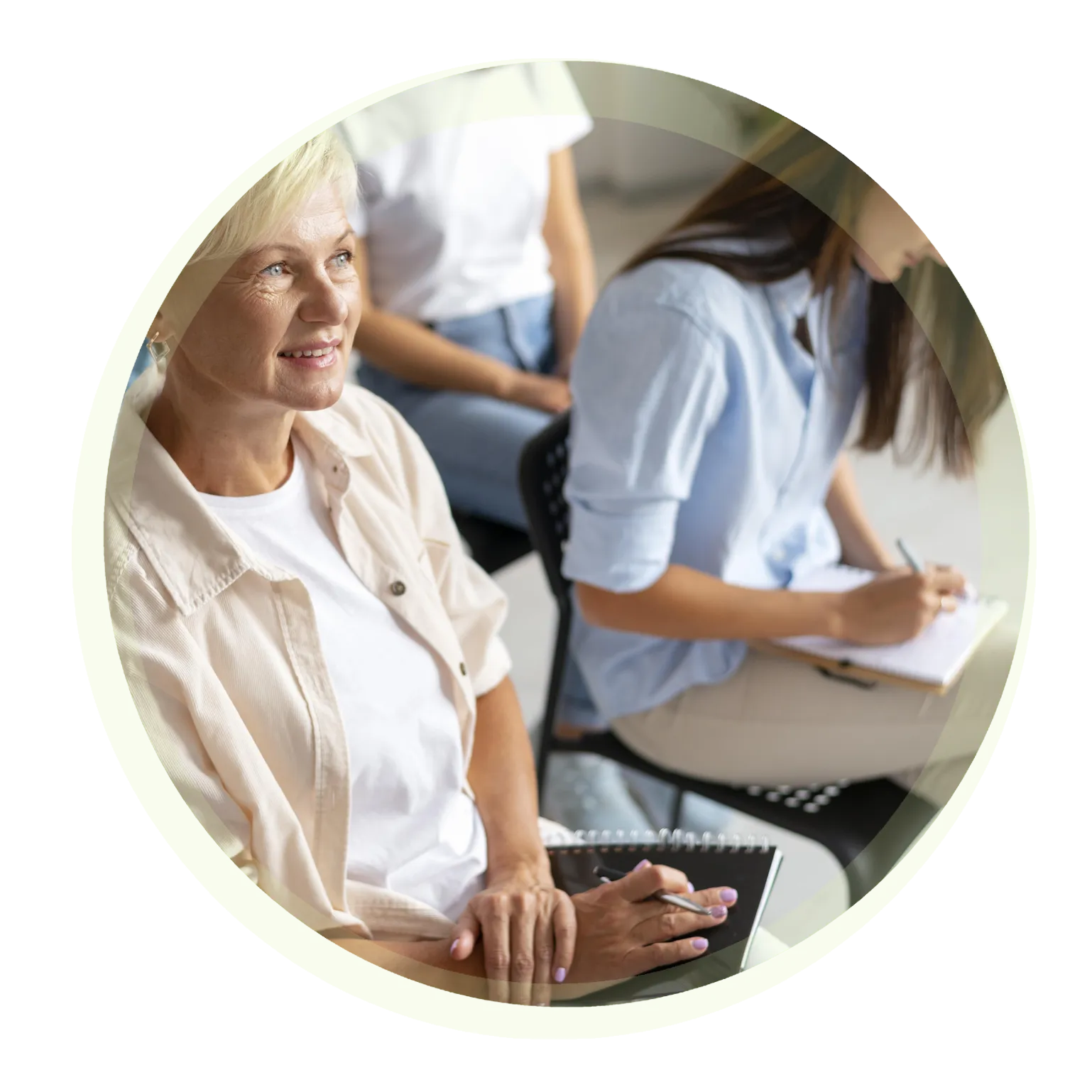 Eine Gruppe von Menschen sitzt in einer Reihe. Im Vordergrund sitzt eine Person mit kurzen, blonden Haaren und einem Notizbuch auf dem Schoß. Daneben schreibt eine Person in einem blauen Hemd in ein Notizbuch.