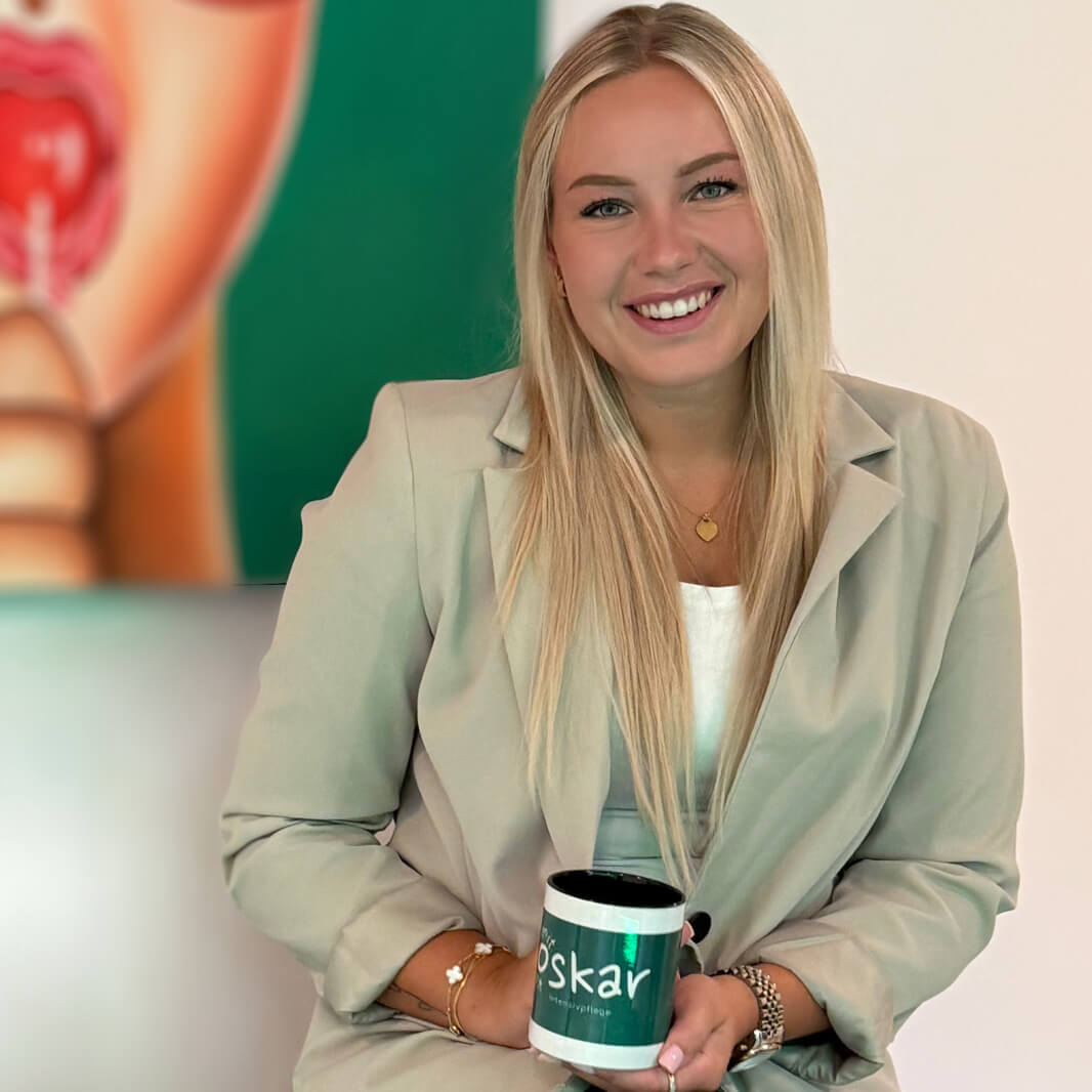 Eine lächelnde Person mit langen, blonden Haaren sitzt vor einem abstrakten Kunstwerk. Die Person trägt einen beigen Blazer und hält eine weiße Tasse mit der Aufschrift "Oskar".