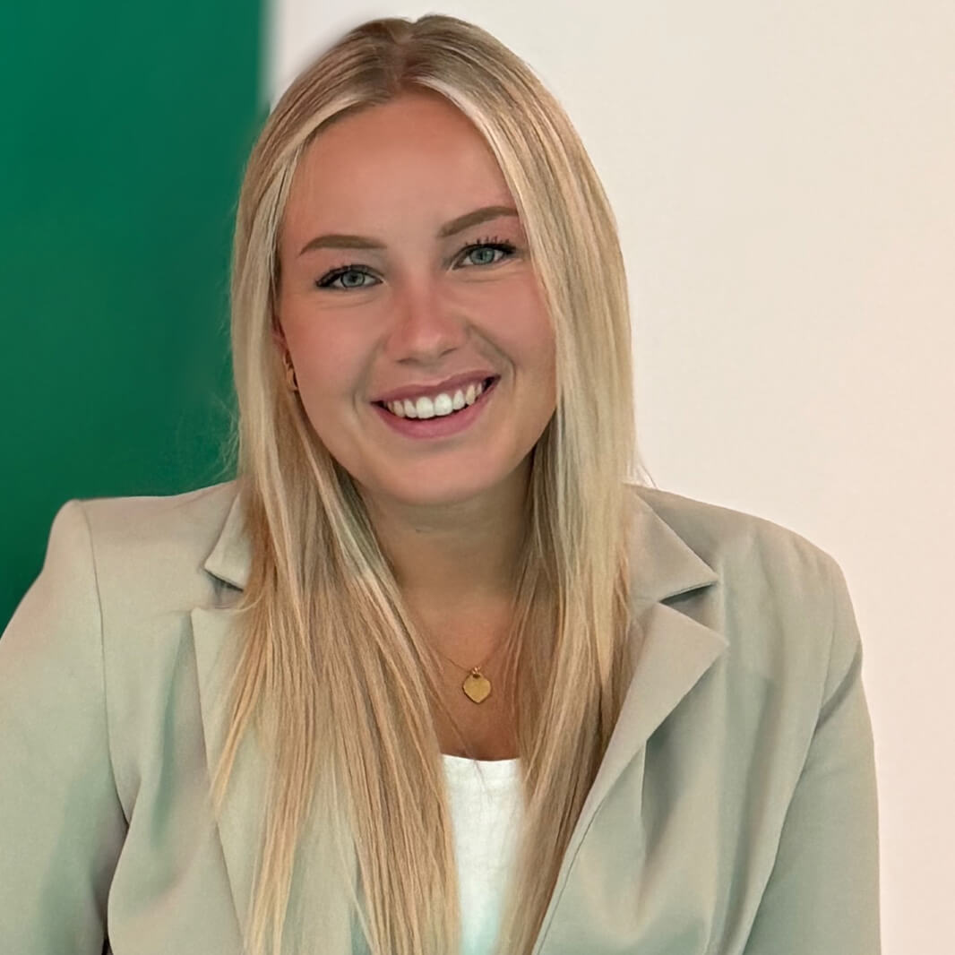 Eine lächelnde Person mit langen blonden Haaren trägt einen hellen Blazer vor einem Hintergrund, der in zwei Hälften geteilt ist: links grün, rechts weiß. Die Person hat ein herzförmiges Medaillon um den Hals.