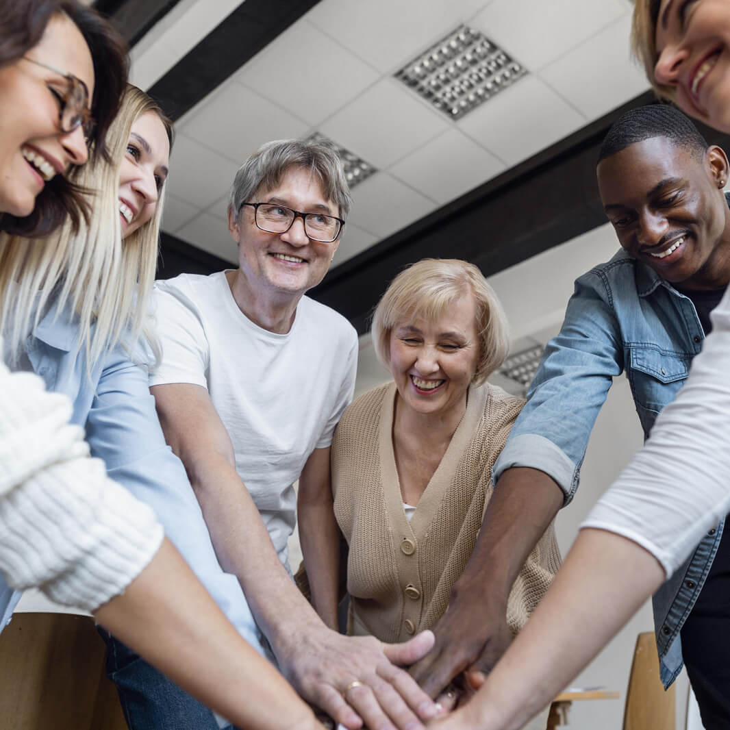 Eine Gruppe von sechs Menschen steht im Kreis und legt die Hände übereinander in der Mitte. Alle lächeln freundlich. Die Umgebung scheint ein heller Raum mit Deckenlampen zu sein.