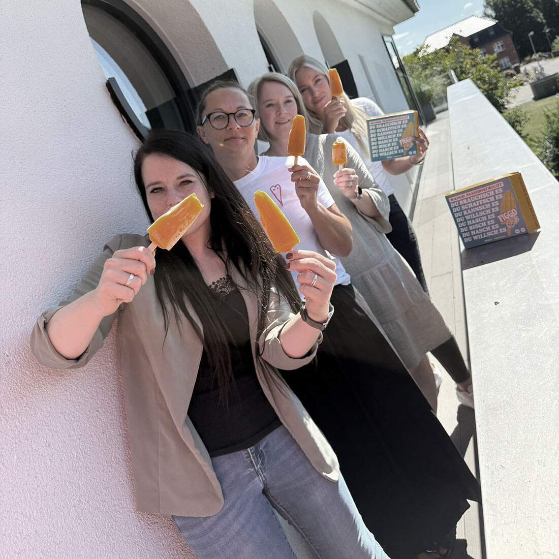 Oskar Intensivpflege Teambild 01 Vier Personen stehen nebeneinander an einer weißen Wand im Freien und halten orangefarbene Eis am Stiel hoch. Sie lächeln und scheinen die kühle Erfrischung zu genießen. Neben ihnen steht eine Box mit Eiswaffeln.