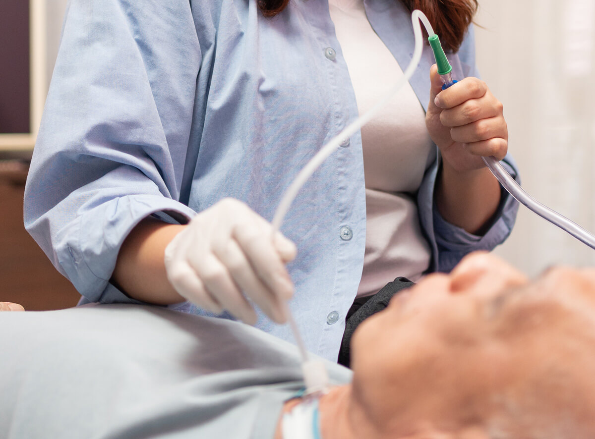 Eine Person in einem hellblauen Kittel und mit weißen Handschuhen hält einen medizinischen Schlauch, der an einen älteren Patienten angeschlossen ist. Der Patient liegt auf einer Liege und schaut zur Decke.
