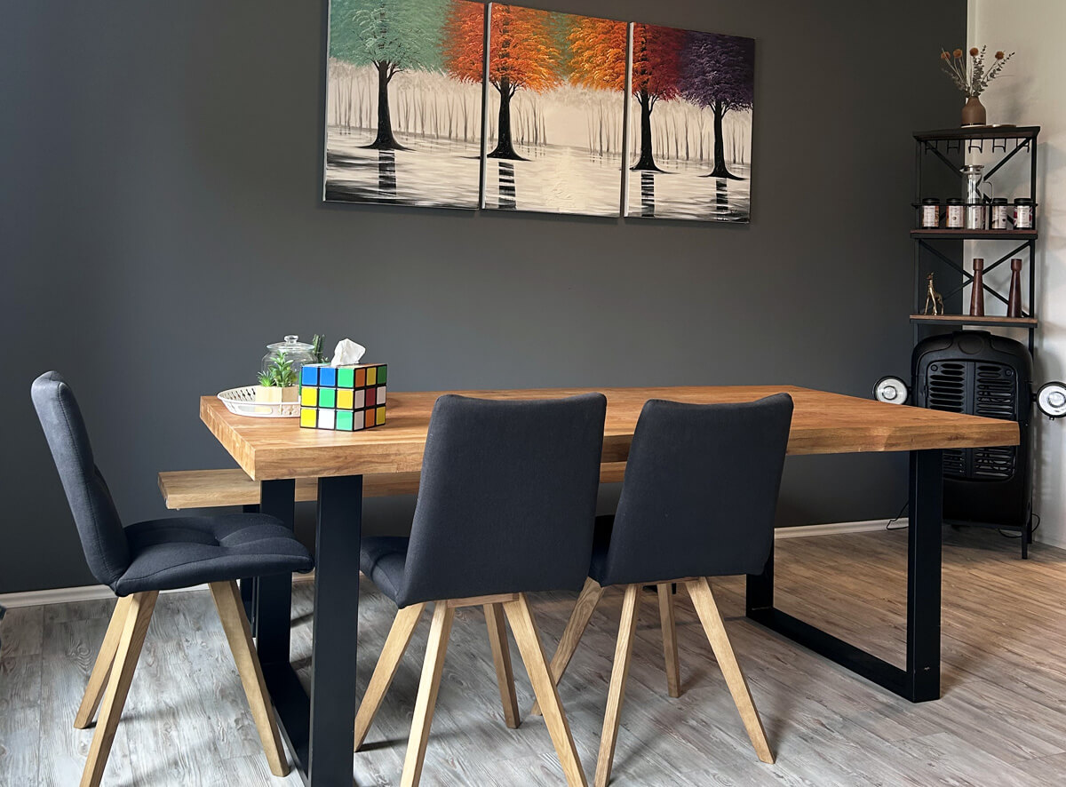 Ein Essbereich mit einem Holztisch und vier dunkel gepolsterten Stühlen. An der Wand hängt ein buntes Gemälde mit Bäumen. Neben dem Tisch steht ein Regal und eine Heizung. Auf dem Tisch liegt ein Rubik's Cube.