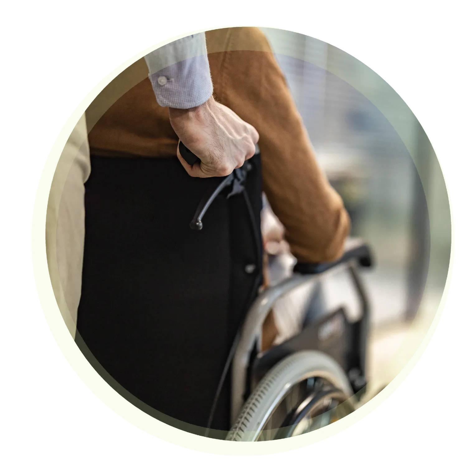 Eine Person schiebt eine andere Person in einem Rollstuhl. Der Fokus liegt auf der Hand des schiebenden Menschen mit leicht unscharfem Hintergrund. Der Rahmen des Bildes ist grünlich rund.