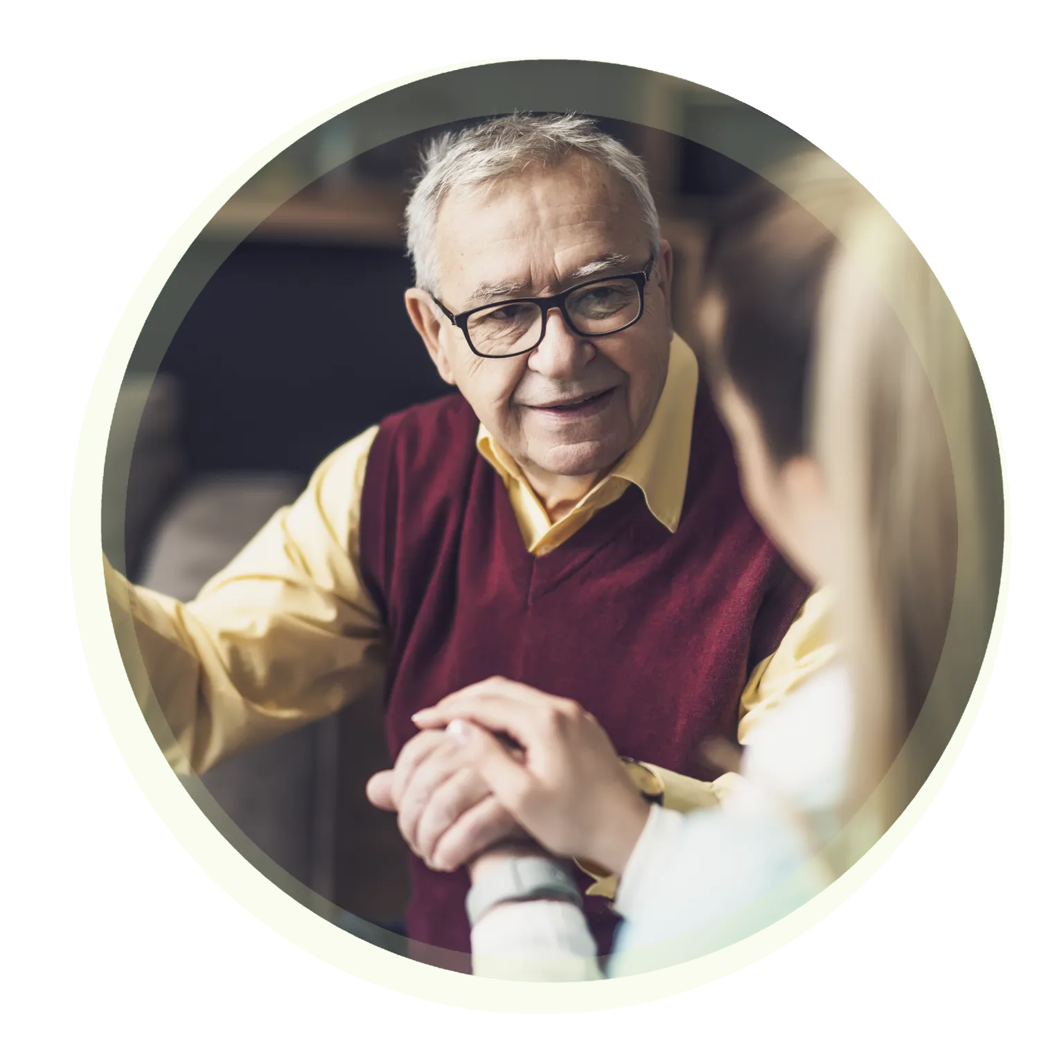 Ein älterer Mann mit Brille und rotem Pullunder sitzt und hält liebevoll die Hand einer sitzenden Person. Beide lächeln. Die Szene wirkt warm und freundlich.